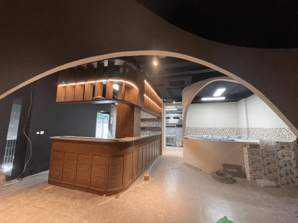 Ảnh thi công thực tế – quầy bar sushi gỗ óc chó đang hoàn thiện, hệ vòm cuốn thạch cao tạo hình hữu cơ đặc trưng, khu bếp ốp gạch mosaic phía sau