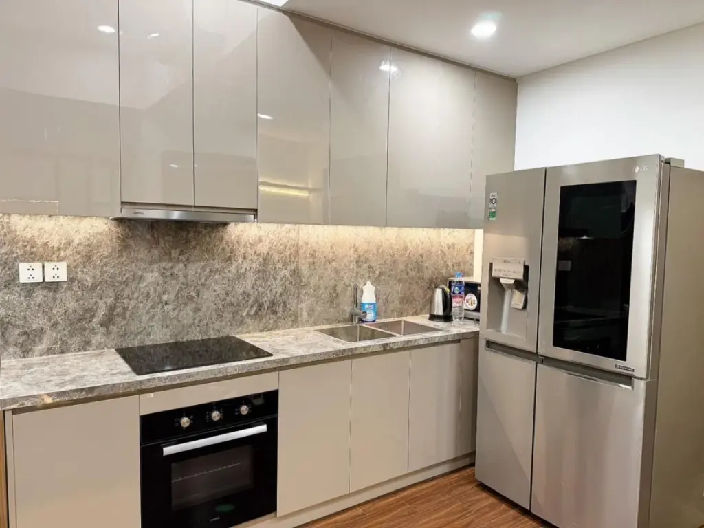 Ảnh thực tế – bếp chữ L cánh bóng màu be, đá ốp vân nâu, lò nướng âm tủ, hốc tủ lạnh