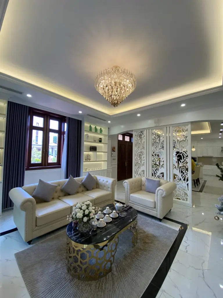 Ảnh thực tế – phòng khách sofa da kem, vách CNC hoa văn trắng, đèn chùm pha lê
