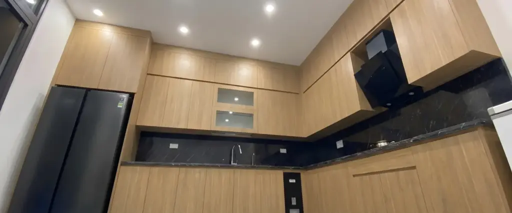 Ảnh thực tế – bếp chữ L cánh gỗ Melamine màu gỗ sáng trên thùng inox 304, khoang tủ lạnh âm tủ